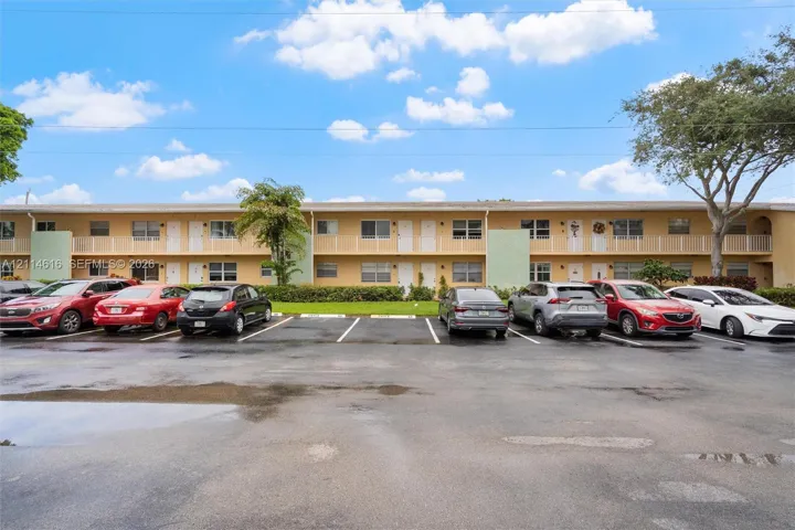 Two-story exterior featuring tan stucco siding, white balcony railings, and a paved parking lot with designated spaces
