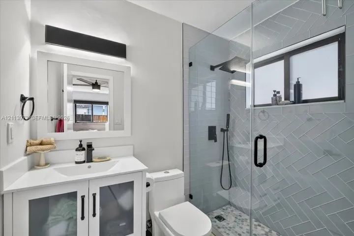 White vanity with a rectangular sink, herringbone-patterned wall tile, glass-enclosed shower with matte black fixtures, and custom light fixture
