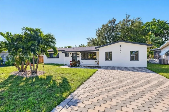 Single-story exterior featuring a light-toned smooth facade, concrete paver driveway, dark-framed windows, and a manicured front lawn with established palms