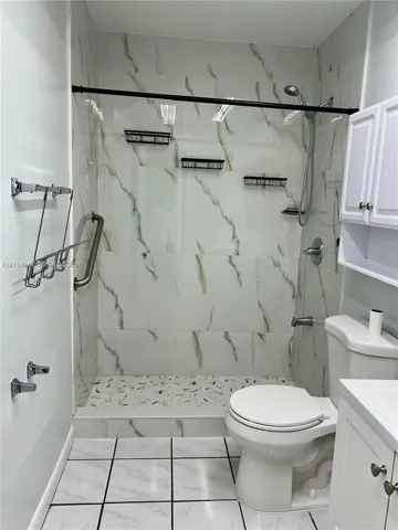 Walk-in shower featuring large-format wall tile with veining, a textured stone-finish floor, wall-mounted grab bar, and adjustable handheld shower head, accompanied by a white vanity with built-in upper cabinetry