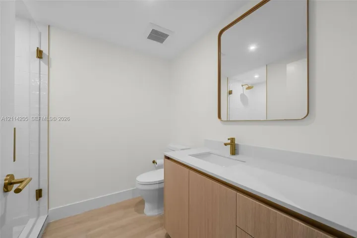 Guest full bathroom featuring a wood-finish vanity, solid surface countertop, brass-toned faucet and hardware, glass-enclosed shower with tiled surround, and a rectangular mirror with a matching metallic frame
