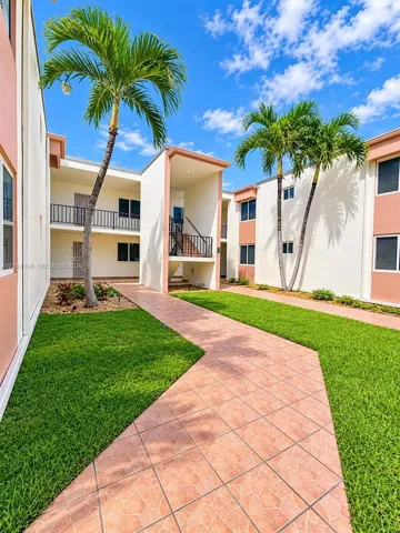 Exterior courtyard featuring a terracotta-tiled walkway, manicured lawn, palm trees, and multi-level buildings with metal railings