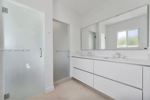 Bathroom featuring a dual-sink floating vanity with white cabinetry, a full-width wall mirror, oversized tile flooring, and frosted glass door enclosures
