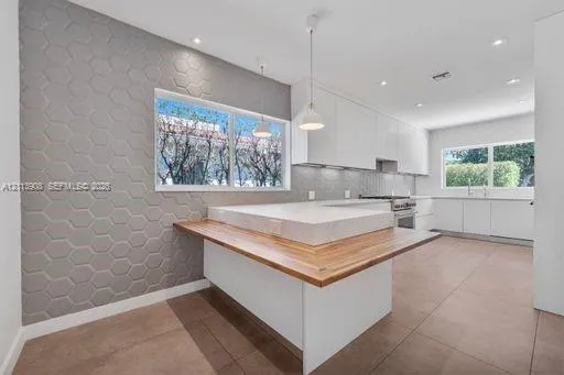 Kitchen featuring matte white flat-panel cabinetry, hexagonal-patterned accent wall, stone-topped island with wood-finished breakfast bar, and tile flooring