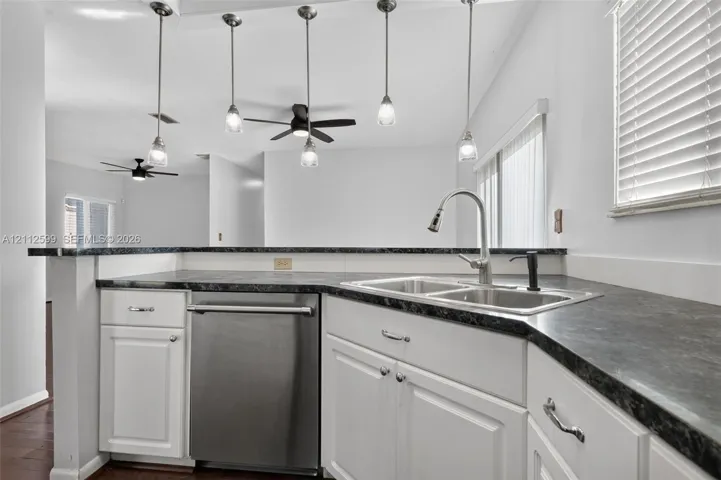 Kitchen featuring white cabinetry, dark stone-finish countertops, stainless steel dishwasher, dual-basin sink with pull-down faucet, and four glass pendant lights