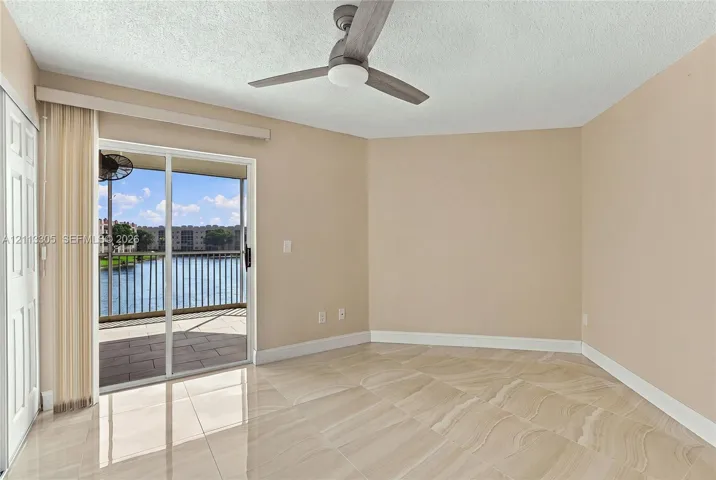 Neutral-toned interior space featuring polished tile flooring, ceiling fan with integrated lighting, and glass sliding doors opening to a screened balcony with water features