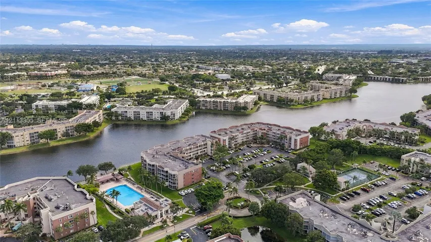 Aerial perspective of a multi-story residential complex featuring waterfront positioning, a swimming pool, tennis courts, and ample surface parking