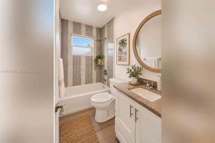 Bathroom featuring a white vanity with a speckled countertop, integrated sink, vertical-tile tub surround, and wood-finish flooring