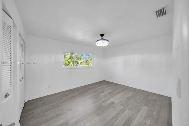 Room featuring wood-finish flooring, white walls, dual louvered-panel closet doors, a central flush-mount light fixture, and a window with exterior greenery