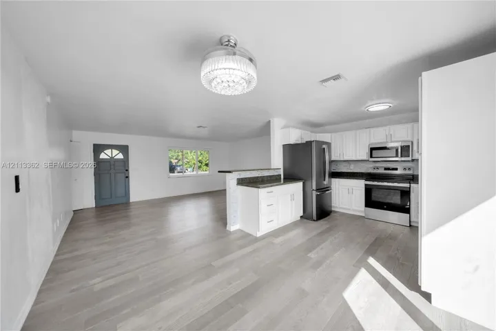 Open-concept kitchen featuring white shaker-style cabinetry, dark stone-finish countertops, stainless steel appliances, and a peninsula island with decorative tile detailing, complemented by wood-finish flooring and a central crystal-style chandelier