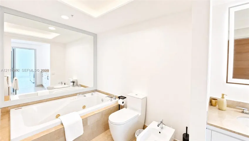 Jetted soaking tub with stone-tiled surround, expansive wall-mounted mirror, dual-piece commode, and bidet
