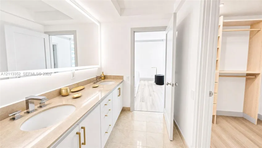 Dual-sink vanity with stone countertop, large illuminated mirror, white cabinetry featuring gold-tone hardware, and an open-concept walk-in closet with built-in shelving