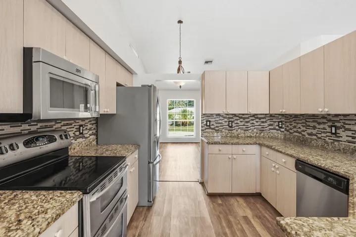 Kitchen featuring light-toned wood cabinetry, stone countertops, stainless steel appliances, multi-textured backsplash, and wood-finish flooring