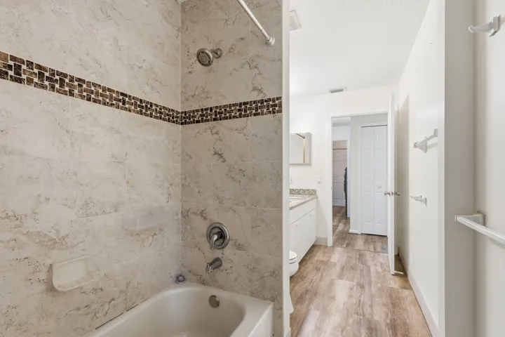 Shower area featuring stone-finish wall tile with mosaic inlay, a chrome shower head and handle, white cabinetry, and wood-finish flooring
