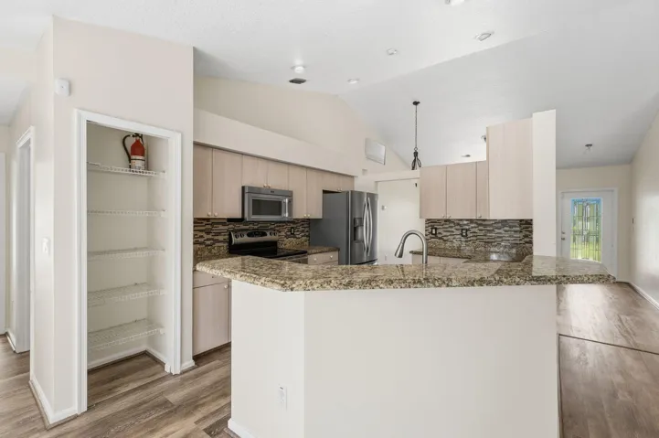 Kitchen featuring a breakfast bar with stone-finish countertops, wood-finish cabinetry, a stacked stone backsplash, stainless steel appliances, and wood-finish flooring under a vaulted ceiling