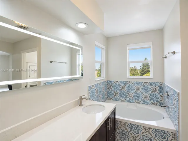 Built-in vanity with white solid-surface countertop and undermount sink, integrated backlit mirror, recessed lighting, and jetted soaking tub featuring patterned ceramic tile surround