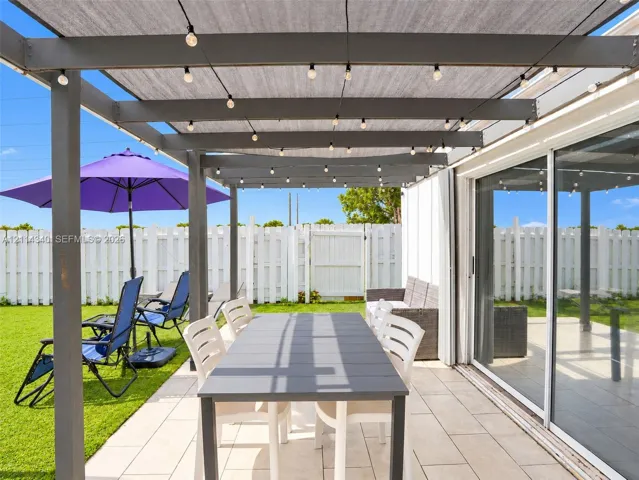 Covered patio featuring a grey wooden pergola, tile flooring, and integrated string lighting, complemented by a white privacy fence