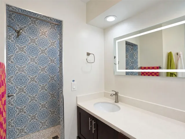 Bathroom featuring a shower stall with patterned tile walls and stone-tiled flooring, dark-finish cabinetry, a solid-surface countertop with an integrated basin, and an illuminated rectangular mirror