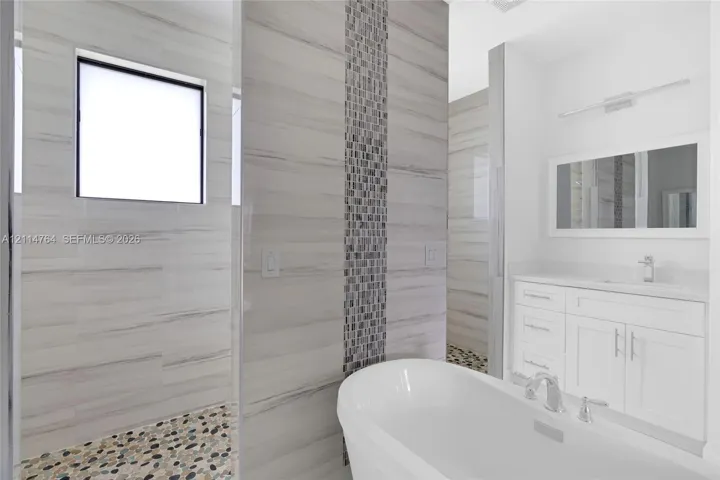 Freestanding soaking tub featuring a chrome faucet, white vanity with recessed-panel cabinetry, stone-tiled shower floor, wall-mounted light fixture, and horizontal-pattern wall tile with a vertical mosaic accent stripe
