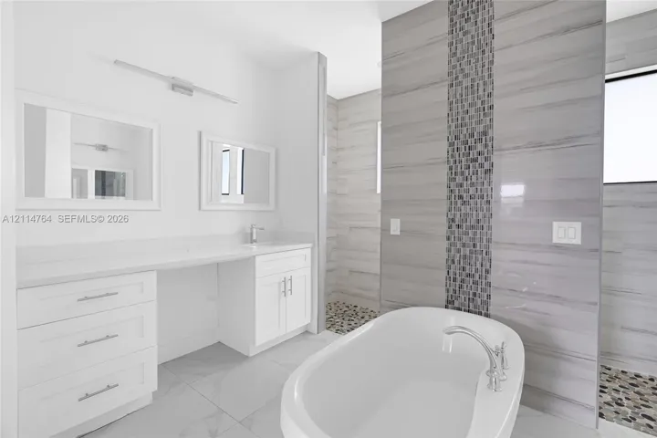 White vanity cabinet with a stone countertop, freestanding soaking tub, chrome finish fixtures, large-format tile flooring, and a walk-in shower with stone tile wall accents