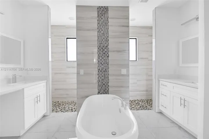 Freestanding soaking tub, dual white vanities with integrated countertops, large-format tile flooring, and a walk-in shower featuring a decorative mosaic tile inlay and stone pebble flooring