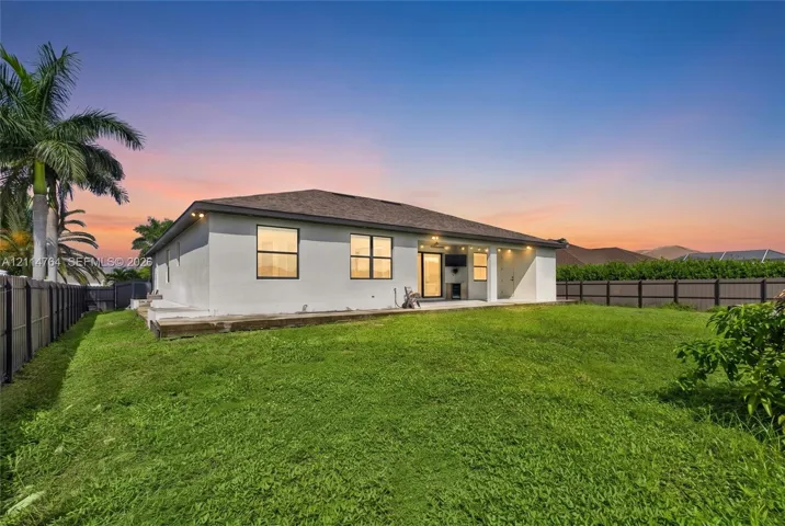 Rear exterior featuring stucco siding, a covered patio, rectangular windows, and a fenced-in grassy yard with mature palm trees