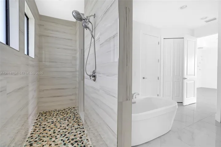 Walk-in shower featuring multi-toned stone pebble flooring, large-format tile surround, and a dual-head chrome shower system, paired with a freestanding soaking tub and polished tile flooring