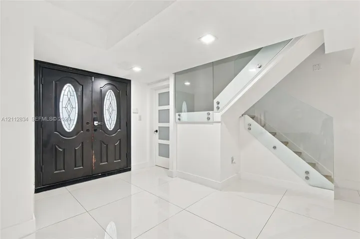 Entryway featuring large-format tile flooring, a double-entry door with oval glass insets, a multi-panel door with frosted glass, and a glass-paneled staircase railing with metal hardware