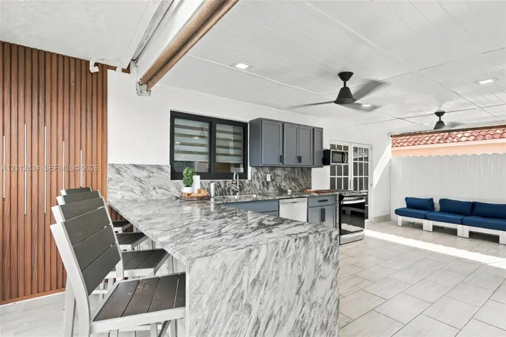 Covered outdoor kitchen featuring a stone-topped peninsula, built-in stainless steel appliances, grey cabinetry, and light-toned floor tiling under a tongue-and-groove ceiling with recessed lighting