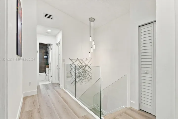 Hallway featuring wood-finish flooring, glass panel railing, multi-pendant light fixture, and louvered bi-fold closet doors