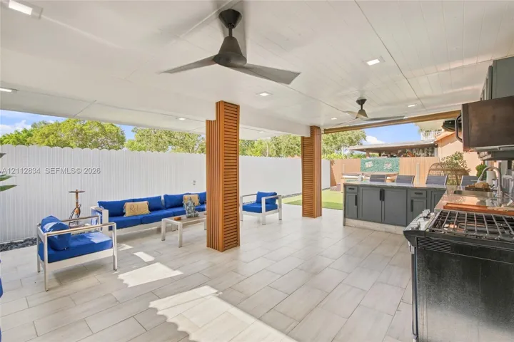 Covered outdoor living area featuring light-toned tile flooring, recessed ceiling lighting, slat-wood wrapped structural columns, and an integrated outdoor kitchen with charcoal cabinetry