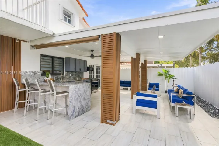 Covered outdoor patio featuring light-toned tile flooring, built-in kitchen island with stone-finish countertop, slatted wood-finish support columns, and recessed lighting