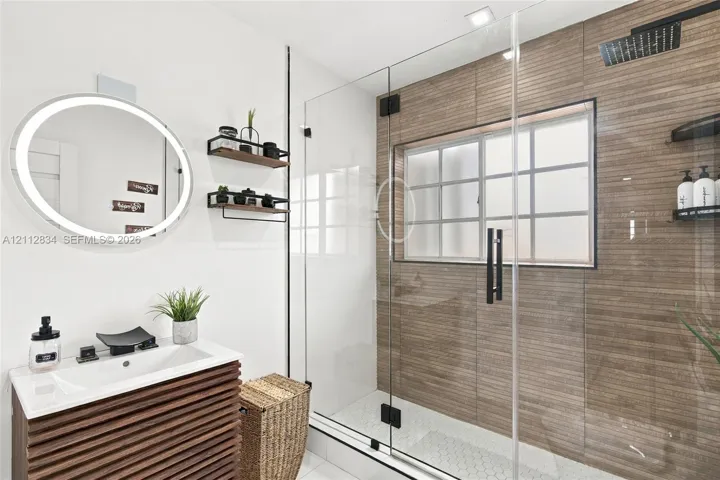 Bathroom featuring a slatted wood-finish vanity, illuminated oval mirror, glass-enclosed shower with horizontal wall tile, mosaic floor tile, and a rainfall showerhead