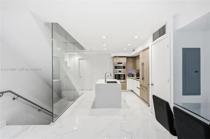 Modern kitchen featuring light wood-finish cabinetry, built-in stainless steel appliances, a central island with a sink, and stone-finish tile flooring