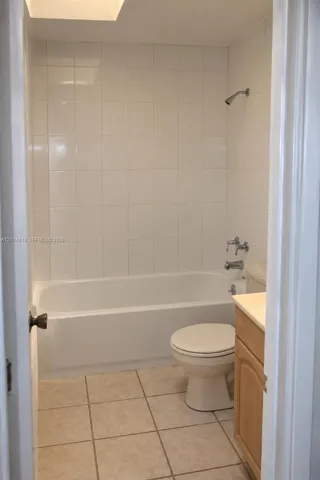 Full-height wall tile surrounding a white tub-shower combination, beige floor tile, and a wood-finish vanity with a countertop sink