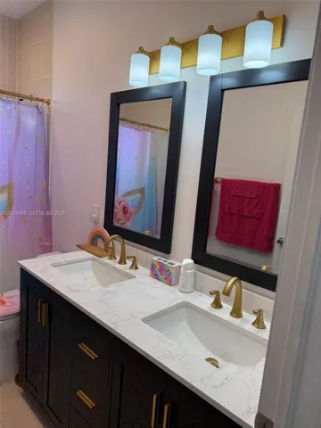 Dual-sink vanity featuring a white stone-pattern countertop, dark finish cabinetry with geometric detailing, brass-toned hardware, dual framed mirrors, and a four-light fixture with cylindrical glass shades