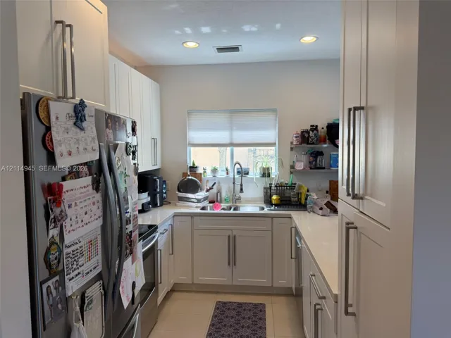 Kitchen featuring white shaker-style cabinetry, solid surface countertops, a stainless steel double-basin sink with a high-arch pull-down faucet, recessed ceiling lighting, and large-format tile flooring