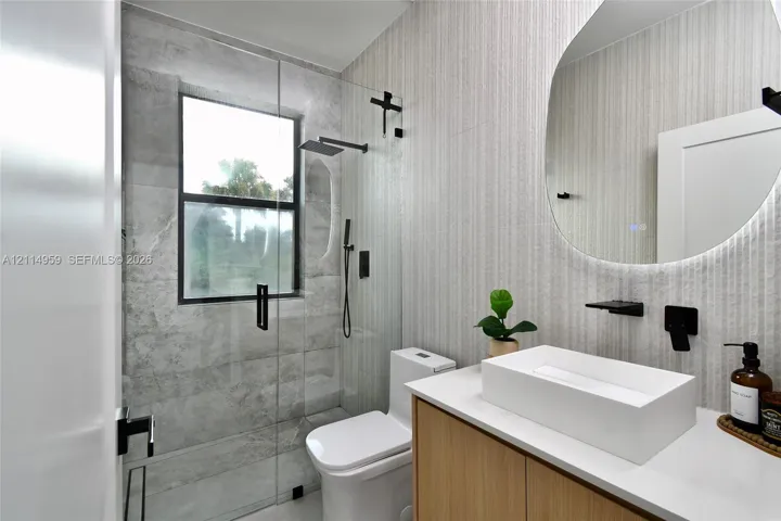 Bathroom featuring a floating wood-finish vanity with a rectangular vessel sink, wall-mounted matte black faucet fixtures, a frameless glass shower enclosure with large-format tile, and an illuminated organic-shaped mirror