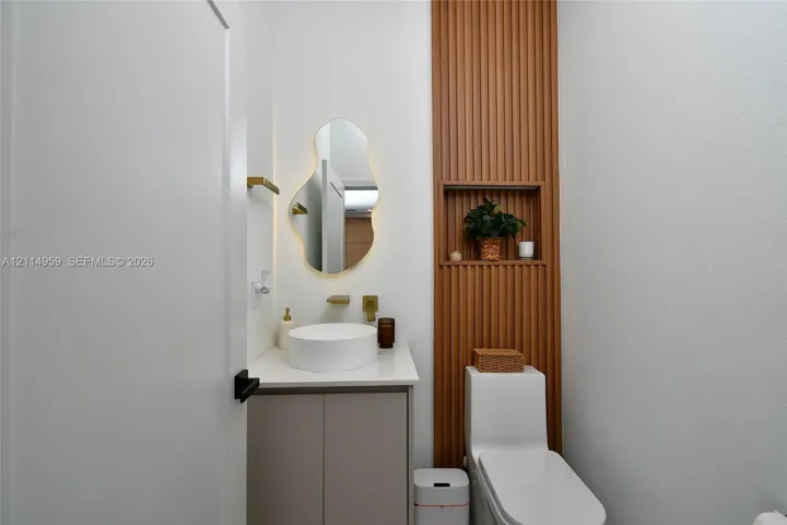 Custom vanity with a solid surface countertop and circular vessel sink, wall-mounted metallic faucet, asymmetrical backlit mirror, and vertical wood-slat accent wall featuring a recessed built-in niche