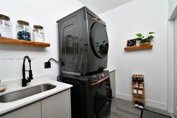 Stacked laundry appliances featuring a metallic finish, undermount stainless steel sink, matte black gooseneck faucet, quartz-surfaced countertop, and natural wood-finish wall-mounted shelving