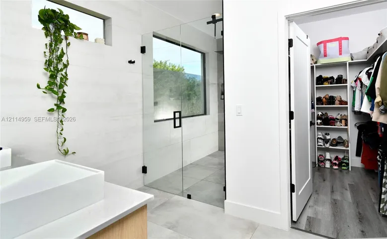 Bathroom featuring a frameless glass-enclosed shower with matte black hardware, large-format tile flooring, rectangular vessel sink on a stone-topped vanity, and a walk-in closet with built-in shelving and wood-finish flooring