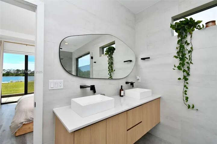Floating wood-finish vanity with a white countertop and dual rectangular vessel sinks, complemented by matte black wall-mounted faucets and an asymmetrical vanity mirror