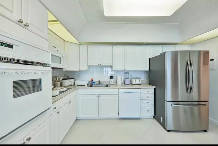 Kitchen featuring white shaker-style cabinetry, solid-surface countertops, recessed ceiling lighting, a stainless steel refrigerator, and built-in wall oven