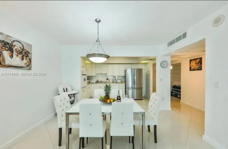 Dining area featuring large-format ceramic tile flooring and a polished metal pendant light fixture, extending to a kitchen with white cabinetry and stainless steel refrigerator