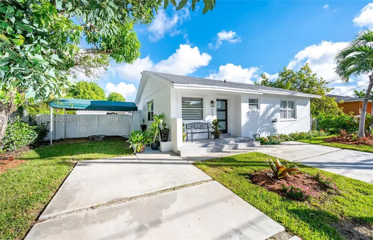 Single-story exterior featuring a painted stucco finish, concrete driveway, front entrance with a modern multi-panel door, metal awning, and privacy fencing