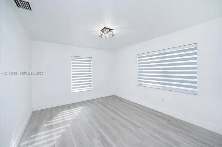 Neutral-toned wood-finish flooring, two windows featuring dual-layer zebra roller shades, and a geometric flush-mount ceiling light fixture