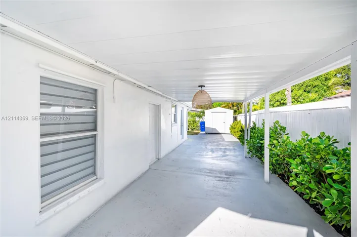 Covered concrete patio featuring a white pergola-style ceiling, dual-pane horizontal blind windows, and a detached exterior storage structure