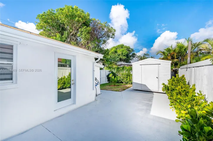 Exterior space featuring a concrete patio, white-painted masonry wall, glass-paneled entry door, detached storage shed, and perimeter fencing
