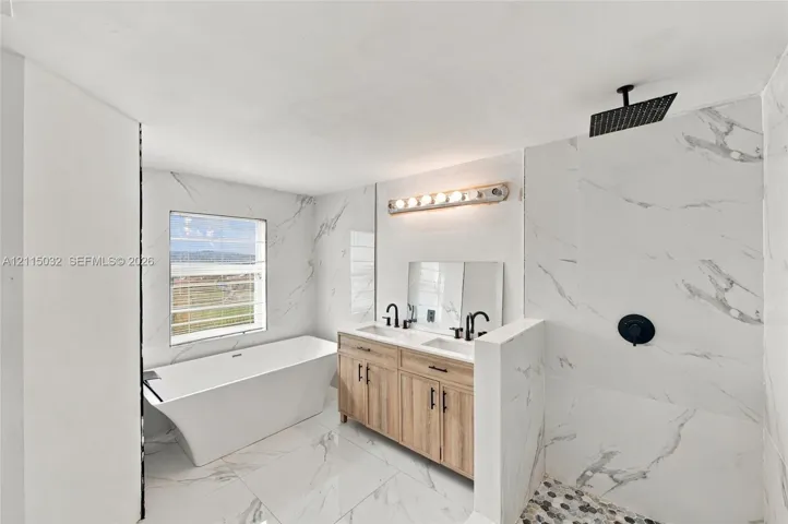 Bathroom featuring a freestanding soaking tub, light wood-finish dual vanity with undermount sinks, stone-patterned tile flooring, full-height wall tiling, and a walk-in shower with a rainfall showerhead