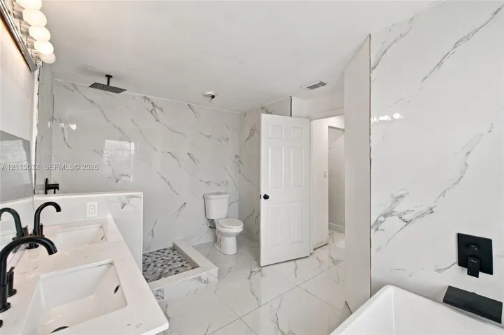 Bathroom featuring large-format stone-patterned wall and floor tiling, dual-sink vanity with white countertop, walk-in shower with stone pebble flooring, and matte black plumbing fixtures